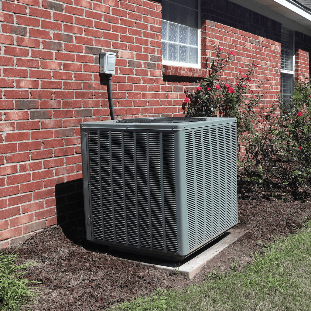 Outdoor air conditioning unit sits on a concrete pad next to a brick house, surrounded by a flower bed and electrical box on the wall.